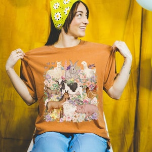 May include: A woman wearing a brown t-shirt with a colorful illustration of farm animals and flowers. The shirt has a relaxed fit and short sleeves. The woman is wearing a green daisy-patterned headband.