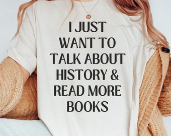 Puede incluir: Camiseta color crema con el texto "I JUST WANT TO TALK ABOUT HISTORY & READ MORE BOOKS" en negro. La camiseta se lleva con una rebeca color canela y vaqueros azul claro. Se ve un collar dorado.