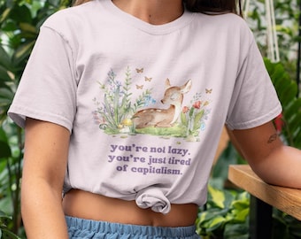 May include: A woman wearing a light purple t-shirt with a graphic of a deer in a field of flowers and the text "you're not lazy, you're just tired of capitalism." She is sitting on a wooden bench with a green smoothie in front of her.