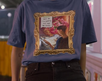 Könnte beinhalten: Ein blaues T-Shirt mit einem gerahmten Bild mit Blumen und einer lesenden Person. Der Text auf dem Shirt lautet "tell me I'm pretty and buy me books". Die Person im Bild trägt schwarze Shorts.