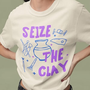 May include: A white t-shirt with a purple graphic that says "Seize the Clay" with a blue clay pot and other pottery tools.