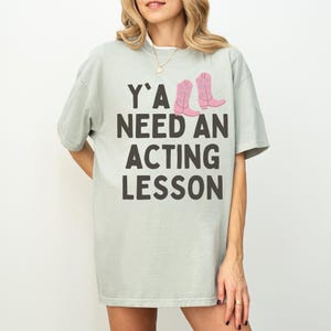 Może przedstawiać: Jasnozielona koszulka z napisem "Y'ALL NEED AN ACTING LESSON" w czarnych literach. Dwa różowe kowbojskie buty są nadrukowane nad słowem "LESSON". Koszulka ma luźny krój.