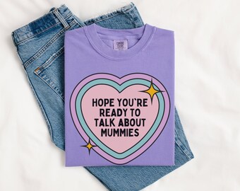 May include: A lilac t-shirt featuring a heart-shaped design with the text "HOPE YOU'RE READY TO TALK ABOUT MUMMIES." The heart has pink, blue, and black outlines with yellow starbursts. The shirt is folded on blue denim jeans.