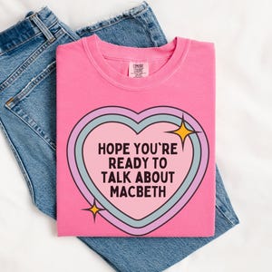 May include: A pink crew neck t-shirt with the text "HOPE YOU'RE READY TO TALK ABOUT MACBETH" inside a heart-shaped design. The shirt is folded on top of a pair of blue jeans.