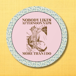 May include: Round coaster with a light pink center featuring a vintage illustration of a king and the text "NOBODY LIKES AFTERNOON NAPS MORE THAN I DO." The coaster has a floral patterned border in shades of blue and green.