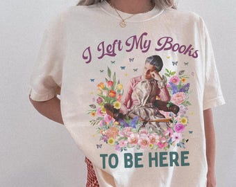 Könnte beinhalten: Cremefarbenes T-Shirt mit einem Blumengrafik und dem Text "I Left My Books TO BE HERE". Das Design zeigt eine Person, die von Blumen und Schmetterlingen in Rosa-, Gelb- und Blautönen umgeben ist. Der Text ist in Lila und Grün gehalten.