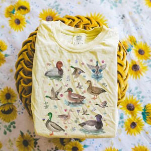 May include: A light yellow t-shirt with a watercolor print of various ducks and flowers. The shirt has a crew neck and short sleeves. The text "COMFORT COLORS" is printed on the tag.