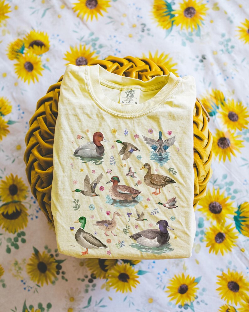 May include: A light yellow t-shirt with a watercolor print of various ducks and flowers. The shirt has a crew neck and short sleeves. The text "COMFORT COLORS" is printed on the tag.