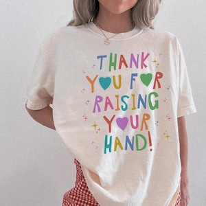 May include: Cream-colored t-shirt with the text "THANK YOU FOR RAISING YOUR HAND!" in colorful, playful lettering. The design includes hearts and starburst accents, creating a cheerful and positive vibe. A casual, comfortable style.