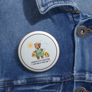 Può includere: Un bottone rotondo con sfondo blu chiaro e bordo a pois bianchi. Il bottone presenta un'illustrazione di un orso bruno che indossa un vestito verde e tiene un cestino di fiori. Il testo sul bottone recita "Attualmente non si accettano vibrazioni negative".