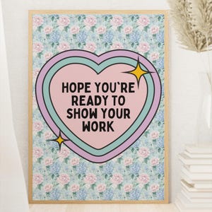 May include: A framed print with a floral background and a heart-shaped graphic. The heart contains the text "HOPE YOU'RE READY TO SHOW YOUR WORK" in a bold font. The frame is light brown.