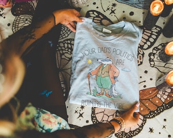 May include: A light blue t-shirt with a cartoon badger wearing a suit and holding a cane. The shirt has the text "Your Dad's Politics Are Ruining The Vibe" printed on it.