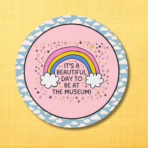 May include: Circular sticker with a light pink backdrop, a rainbow, clouds, and stars. The text reads "It's a beautiful day to be at the museum!" The border is light blue with white clouds.