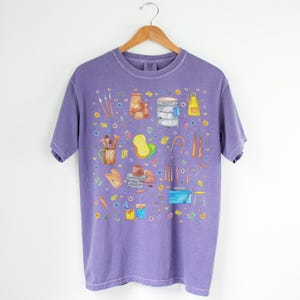 Puede incluir: Una camiseta lila con un diseño de acuarela de herramientas y equipos de cerámica. El diseño incluye pinceles, tornos de alfarero, hornos y otros suministros de arte cerámico, con acentos florales.
