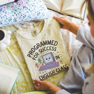 May include: Cream-colored t-shirt with the text "PROGRAMMED FOR SUCCESS IN ENGLISH CLASS" and a cartoon computer graphic. The shirt is displayed on a floral patterned bedspread, with a coffee cup and books nearby.
