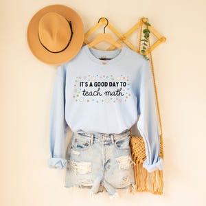 May include: Light blue sweatshirt with the text "It's a good day to teach math" surrounded by colorful flowers. The sweatshirt is hanging on a wooden rack with a brown hat and a woven bag.