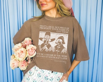 May include: Brown t-shirt with a graphic of two figures in hats and the text "THAT'S A REAL NICE PIECE OF PROPERTY YOUR FAMILY'S GOT THERE. IT'D BE A SHAME IF YOU WERE ACCUSED OF WITCHCRAFT LATER."