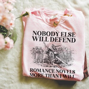 May include: A light pink t-shirt with the text "NOBODY ELSE WILL DEFEND ROMANCE NOVELS MORE THAN I WILL" in black. The shirt features a black and white illustration of a historical battle scene. A gold butterfly necklace rests on the shirt.