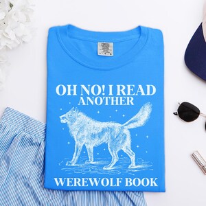 May include: A bright blue t-shirt with the text "OH NO! I READ ANOTHER WEREWOLF BOOK" in white. A white illustration of a wolf is centered below the text. The shirt is folded neatly on a white surface.