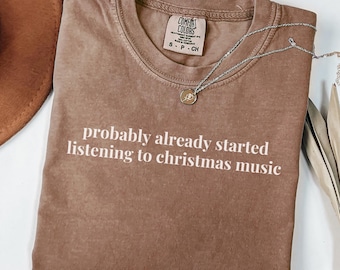 May include: A brown Comfort Colors t-shirt with the text "probably already started listening to christmas music" in white. A silver necklace with a pendant is draped across the shirt. The shirt is folded on a white surface.