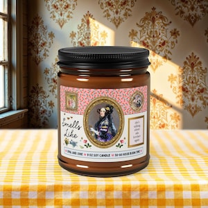 May include: A 9 oz soy candle in a brown glass jar with a black lid. The label features a floral pattern, a portrait of Ada Lovelace in a gold frame, and text that reads: Smells Like, she's talking about ada lovelace again, Opal and June.