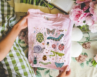 Könnte beinhalten: Pinkes T-Shirt mit einem bunten Insekten- und Schmetterlingsdesign. Das Design beinhaltet eine Schnecke, einen Marienkäfer, eine Gottesanbeterin, eine Raupe und einen Schmetterling.