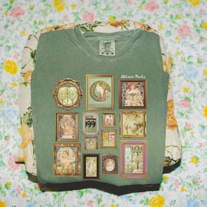 May include: Sage green t-shirt featuring multiple framed art prints. The prints are in gold frames and feature images of women, nature, and art nouveau designs. The text "Alphonse Mucha" is printed on the shirt.