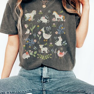 May include: A dark gray t-shirt with a whimsical design featuring a variety of animals and flowers. The animals include a horse, a cow, a duck, a rabbit, a chick, a hedgehog, a mouse, and a bird. The flowers are in shades of pink, blue, and yellow.