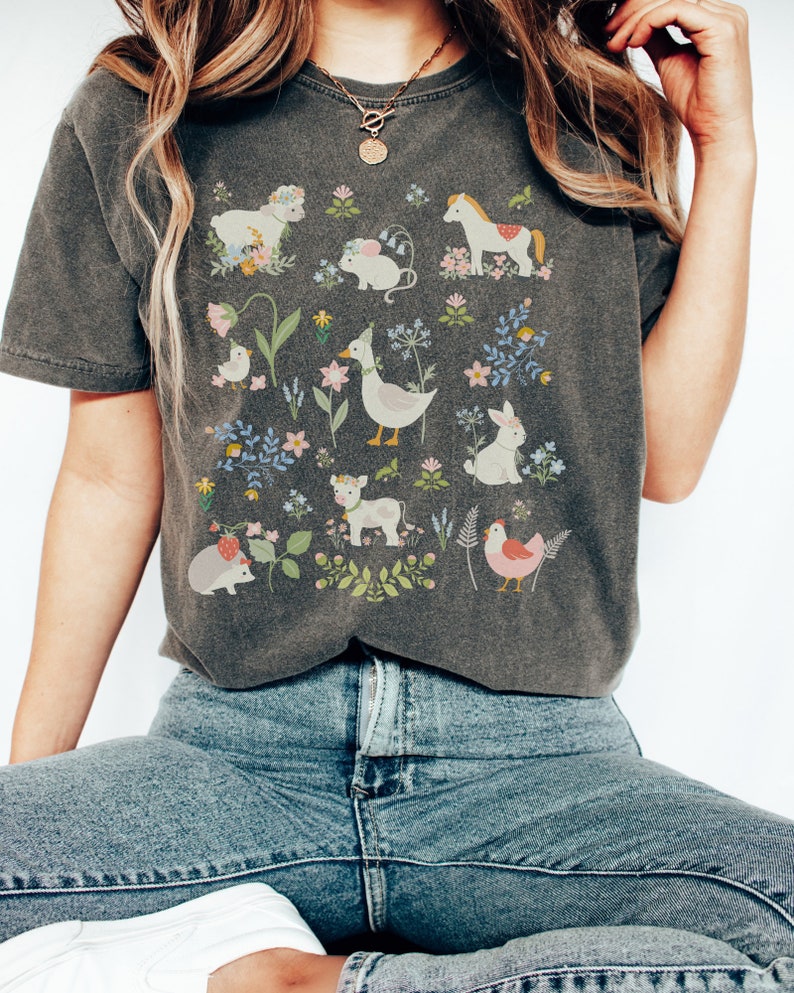 May include: A dark gray t-shirt with a whimsical design featuring a variety of animals and flowers. The animals include a horse, a cow, a duck, a rabbit, a chick, a hedgehog, a mouse, and a bird. The flowers are in shades of pink, blue, and yellow.