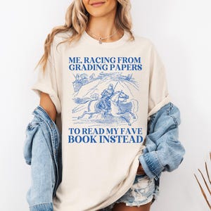 Könnte beinhalten: Cremefarbenes T-Shirt mit blauem Aufdruck eines Reiters zu Pferd und dem Text: „ME, RACING FROM GRADING PAPERS TO READ MY FAVE BOOK INSTEAD“. Das Shirt hat einen lässigen Stil.