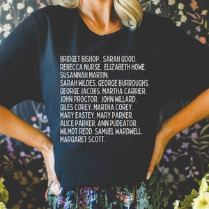 May include: A black short-sleeved t-shirt featuring white text listing the names: BRIDGET BISHOP, SARAH GOOD, REBECCA NURSE, ELIZABETH HOWE, SUSANNAH MARTIN, SARAH WILDES, GEORGE BURROUGHS, GEORGE JACOBS, MARTHA CARRIER, JOHN PROCTOR, JOHN WILLARD, GILES COREY, MARTHA COREY, MARY EASTEY, MARY PARKER, ALICE PARKER, ANN PUDEATOR, WILMOT REDD, SAMUEL WARDWELL, MARGARET SCOTT.