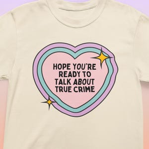 Puede incluir: Una camiseta blanca con un gráfico de corazón rosa que dice "Espero que estés listo para hablar de crímenes reales". El corazón tiene un contorno azul y morado y dos estrellas amarillas.