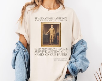Könnte beinhalten: Cremefarbenes T-Shirt mit einem gerahmten Porträt von Alexander Hamilton und Text. Der Text lautet: „Wenn Alexander Hamilton 51 Aufsätze überleben kann, können wir alle in sechs Monaten überleben, unsere Namen auf unsere Papiere zu schreiben.“
