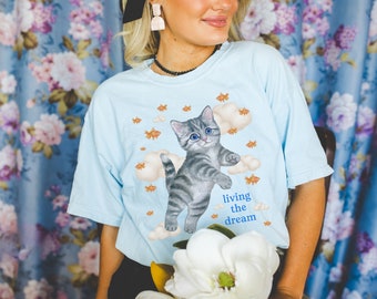 Op de afbeelding: Een lichtblauw T-shirt met een cartoonillustratie van een grijze tabbykat met blauwe ogen, omringd door wolken en oranje vissen. De tekst "living the dream" is onder de kat gedrukt.