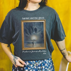May include: Dark gray t-shirt featuring the text "hamlet and the ghost" above a framed painting. The painting depicts a figure on a beach with a ghostly figure. Below the painting is "Frederic James Shields 1901".