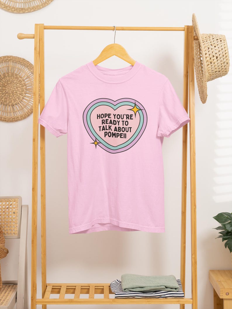 Op de afbeelding: Lichtroze t-shirt met de tekst "HOPE YOU'RE READY TO TALK ABOUT POMPEII" in een hartvormig ontwerp. Het ontwerp heeft een turquoise, lichtblauwe en paarse omtrek met gele steraccenten. Het shirt hangt aan een houten kledingrek.