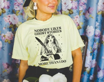 Op de afbeelding: Lichtgele t-shirt met de tekst "NOBODY LIKES GHOST STORIES MORE THAN I DO" en een zwarte illustratie van een vrouw die met een veer schrijft. Het shirt wordt gecombineerd met een zwarte broek.