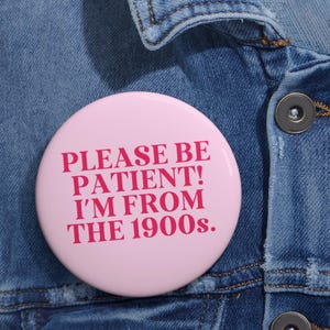 May include: A light pink button with the text "PLEASE BE PATIENT! I'M FROM THE 1900s." in a bold, dark pink font. The button is pinned to a blue denim jacket.