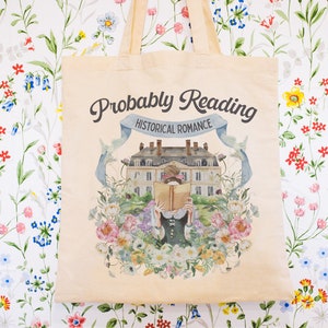 May include: A beige tote bag with a floral design and the text "Probably Reading Historical Romance". The design features a woman reading a book in front of a building with a blue ribbon.