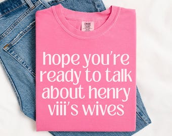 Może przedstawiać: Różowa koszulka z białym napisem "hope you're ready to talk about henry VIII's wives". Koszulka jest złożona na niebieskich dżinsach.