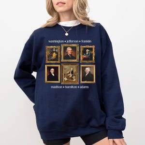 May include: Navy blue sweatshirt featuring six framed portraits of historical figures. The text "washington + jefferson + franklin" and "madison + hamilton + adams" are printed above and below the portraits. The portraits are in gold frames.