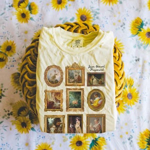 May include: A light yellow t-shirt featuring a print of nine framed paintings. The paintings are all in a similar style and depict people in various poses. The text "Jean-Honor&eacute; Fragonard" is printed below the paintings.