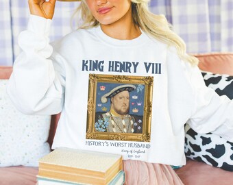 Könnte beinhalten: Weißer Pullover mit einem Porträt von König Heinrich VIII. in einem goldenen Rahmen. Der Text "King Henry VIII" ist in Blau über dem Porträt gedruckt. Unter dem Porträt steht der Text "History's Worst Husband King of England 1509-1547".