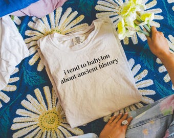 Puede incluir: Una camiseta blanca con el texto "i tend to babylon about ancient history" impreso en ella. La camiseta está sobre una manta de estampado floral azul y blanco.