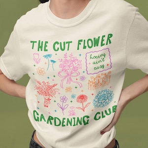 K&ouml;nnte beinhalten: Ein wei&szlig;es T-Shirt mit einer gr&uuml;nen Grafik, auf der "The Cut Flower Gardening Club" steht und ein Blumenstrau&szlig;, ein Blumentopf und der Text "hoeing ain't easy" abgebildet sind.