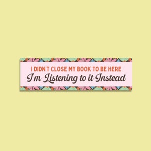 May include: A pink and brown banner with a floral border. The text on the banner reads "I didn't close my book to be here I'm listening to it instead."