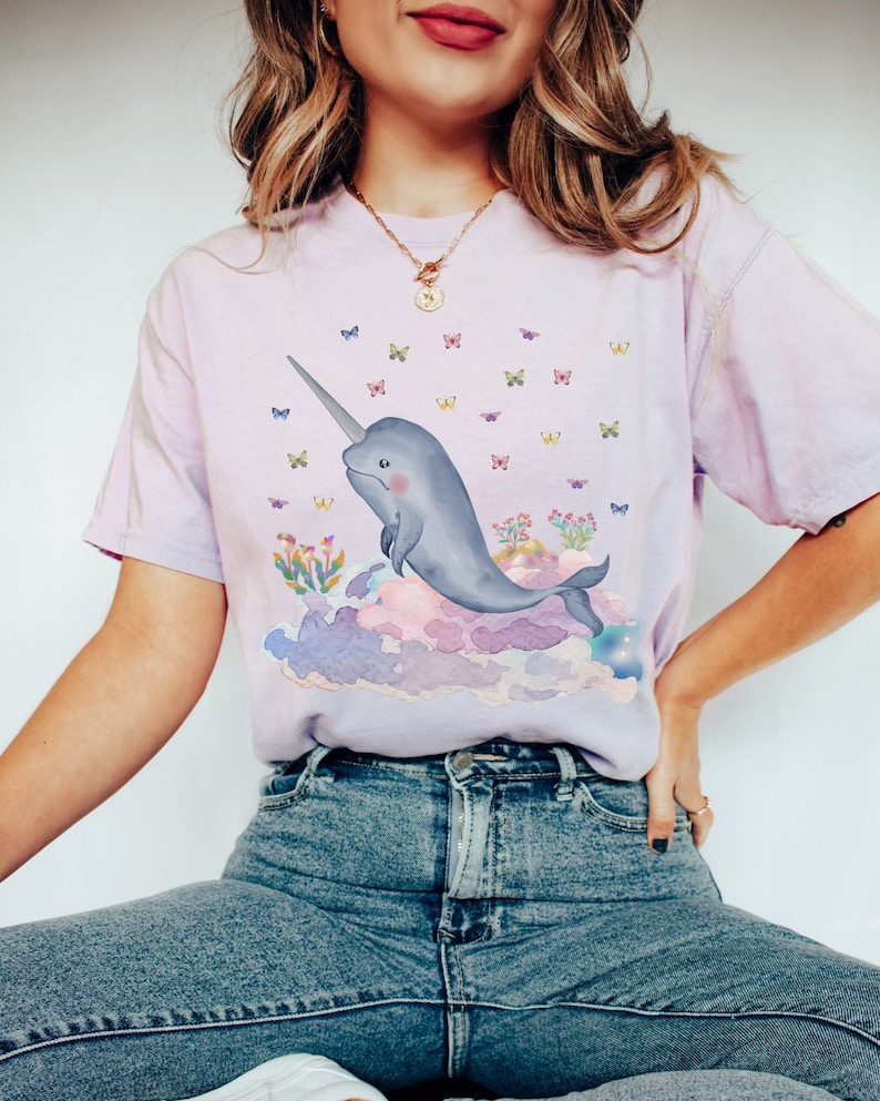 May include: A light purple t-shirt with a watercolor illustration of a narwhal swimming in clouds with butterflies. The narwhal is gray and has a long, spiraled horn.