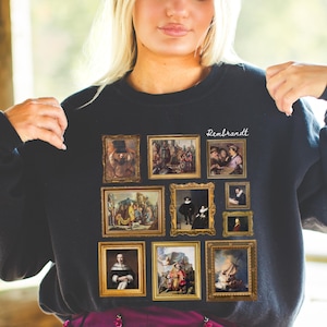 Op de afbeelding: Zwart sweatshirt met een print van verschillende ingelijste schilderijen, waaronder een zelfportret van Rembrandt, en de tekst "Rembrandt" in wit.