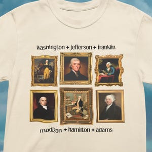 May include: Cream-colored t-shirt featuring portraits of six historical figures in ornate gold frames. The names "Washington + Jefferson + Franklin" and "madison + hamilton + adams" are printed above and below the portraits.