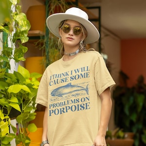 May include: A light beige t-shirt with a blue graphic of a dolphin and the text "I think I will cause some problems on porpoise".